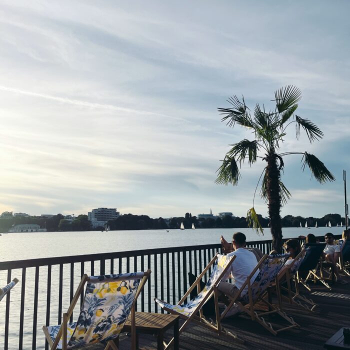 Blick von einem Steg an der Alster mit Außengastro. Liegestühle, eine Palme in einem Kübel, Abendstimmung