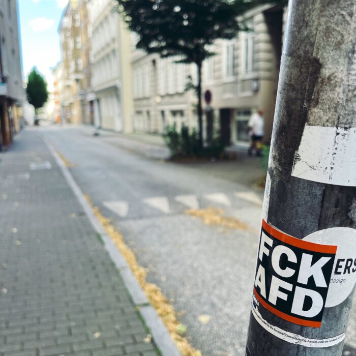 Ein Aufkleber an einem Verkehrsschildmast: "FCKAFD", im Hintergrund unscharf eine leere Straße