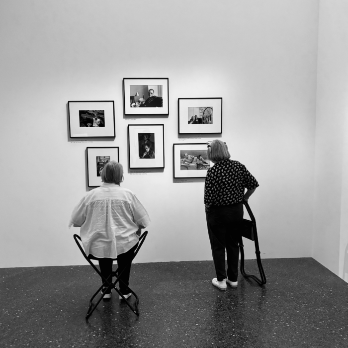 Zwei ältere Damen, von hinten vor Bildern der Cartier-Bresson-Ausstellung fotografiert, eine sitzt auf einem Klappstuhl, die andere hat diesen zusammengeklappt in der Hand. Ein schwarzweißes Bild.