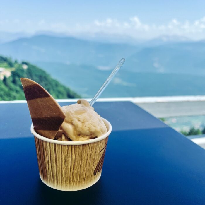 Ein kleiner Eisbecher (Schokolade und Walnuss) vor der Aussicht vom Mendelpass, der Hintergrund ist unscharf