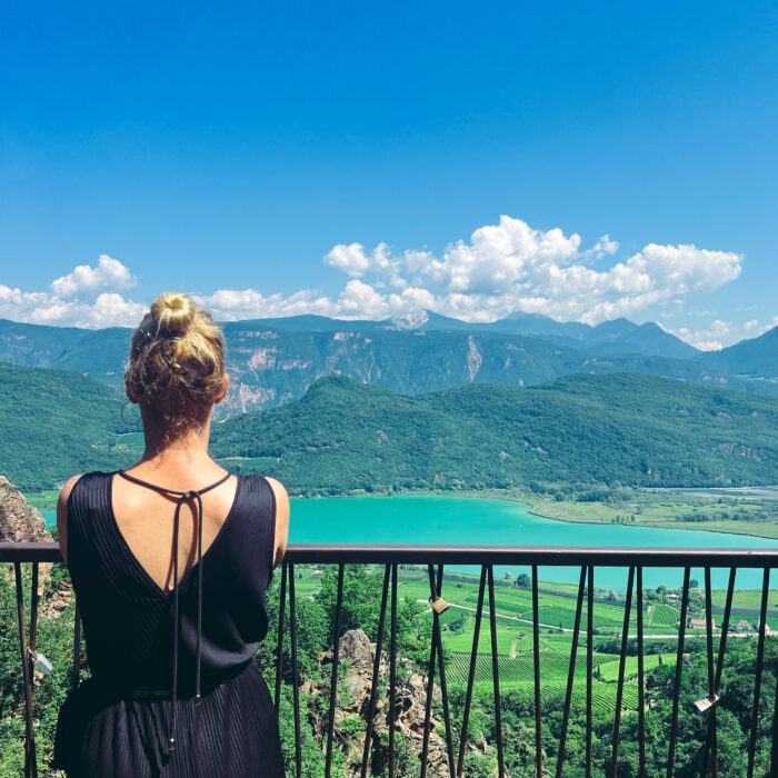 Die Herzdame steht mit dem Rücken zum Fotografen vor einem Geländer an einem Hang, in der Weite, unten, der Kalterer See