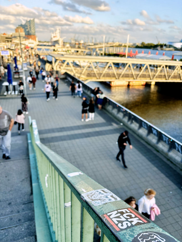 Die Promenade an den Landungsbrücken, von der Treppe aus aufgenommen, die zur S-Bahn-Station führt. Auf dem Treppengeländer ein Aufkleber "FCK NZS", darauf der Fokus, der Hintergrund unscharf