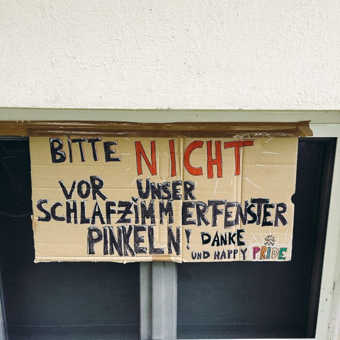 Ein Pappschild an einem Fenster, von Hand beschrieben: "Bitte nicht vor unserem Schlafzimmerfenster pinkeln! Danke und Happy Pride"