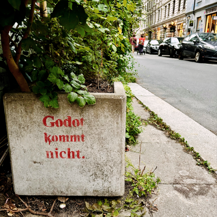 Ein Pflanzkübel aus Beton an einem Straßenrand, in roter Farbe aufgesprüht: "Godot kommt nicht"