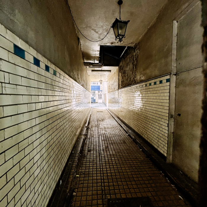 Blick in einen schmalen Gang, der zu einem Hinterhof führt. Gekachelte Wände, in denen sich Licht spiegelt, eine alte Deckenlampe.