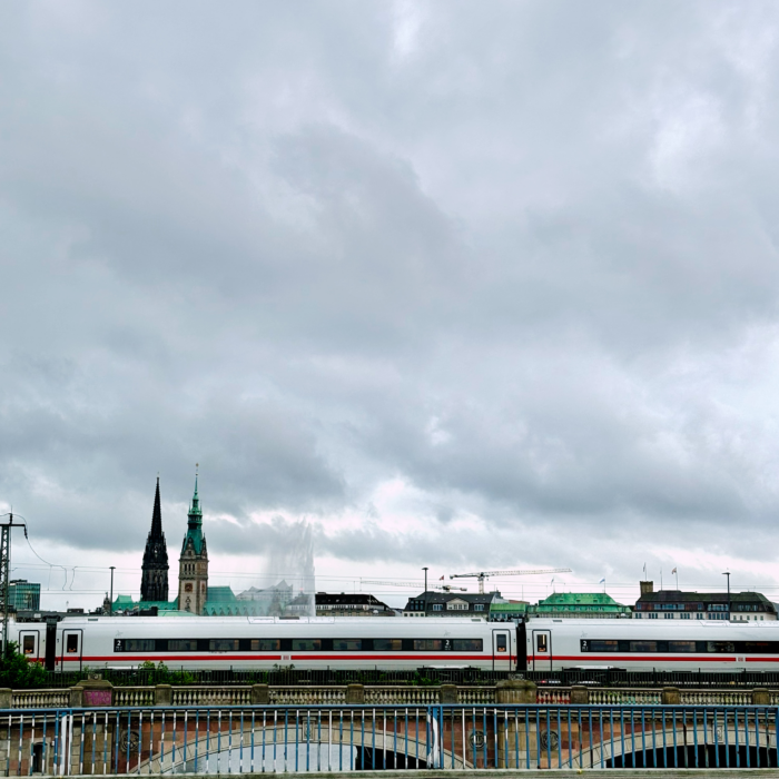 Blick von der Kennedybrücke auf das Rathaus, auf den Gleisen davor ein ICE. Wolkiger Himmel, man erkennt auch die Alsterfontäne.