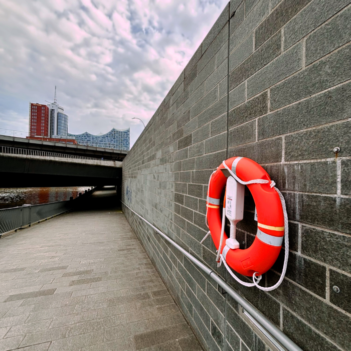 Ein Rettungsring an einer Wand neben dem Alsterfleet, im Hintergrund die Elbphilharmonie, über eine Brücke ragend