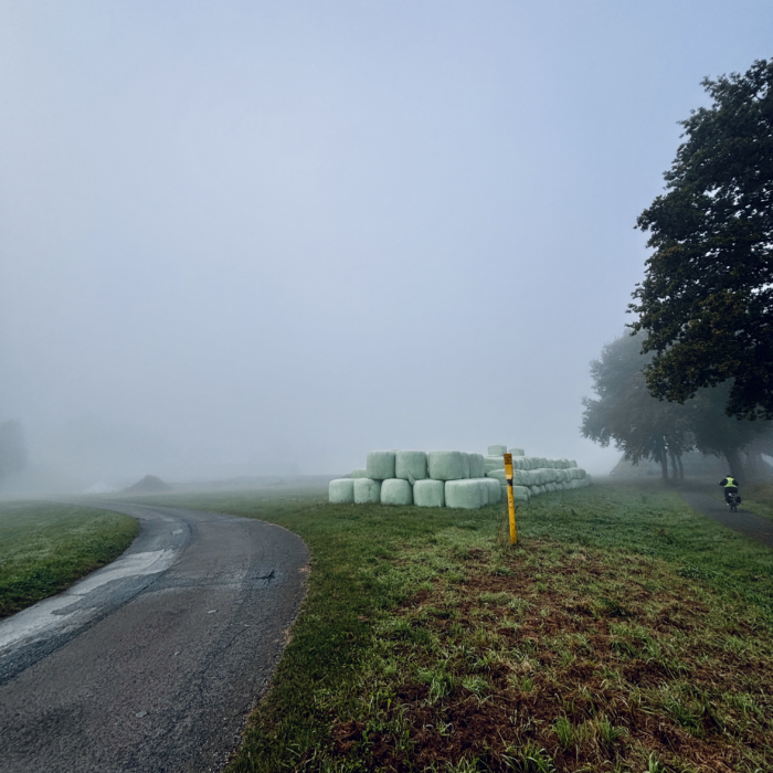 Ein Feldweg, der an eingeschweißten Heuballen vorbeiführt, klein im Bild ein einsamer Radfahrer im Morgennebel