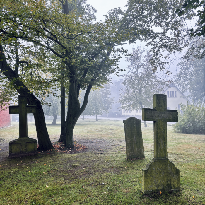 Altes, etwas eingesunkenem, schräg stehende Grabmäler und Kreuze auf einem nebligen Kirchof