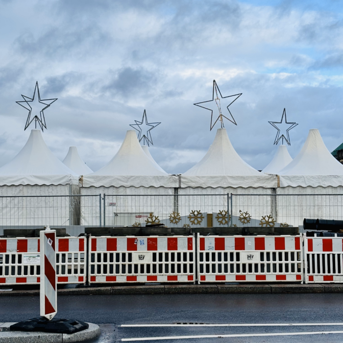 Die weißen Pavillonzelte des Weihnachtsmarktes auf dem Jungfernstieg von der Rückseite, mit großen, unbeleuchteten Sternen auf den Dächern, davor rotweiße Absperrgitter, kein sehr heimeliger Gesamteindruck