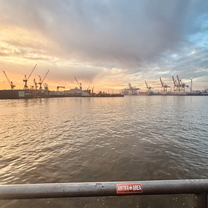 Das Bild zeigt den Hamburger Hafen bei Sonnenaufgang. Im Vordergrund ist ein Geländer mit einem Aufkleber, auf dem 'ANTIFA AREA' steht. Im Hintergrund sind zahlreiche Kräne und industrielle Strukturen zu sehen, die sich entlang des Wassers erstrecken. Der Himmel ist teils bewölkt, und die Sonne erzeugt ein warmes Licht, das sich im Wasser spiegelt.