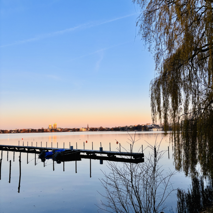 Die Außenalster im Winternachmittagssonnenlicht, im Vordergrund ein Bootsanleger, an dem nur noch ein Segelboot liegt, unter einer Plane