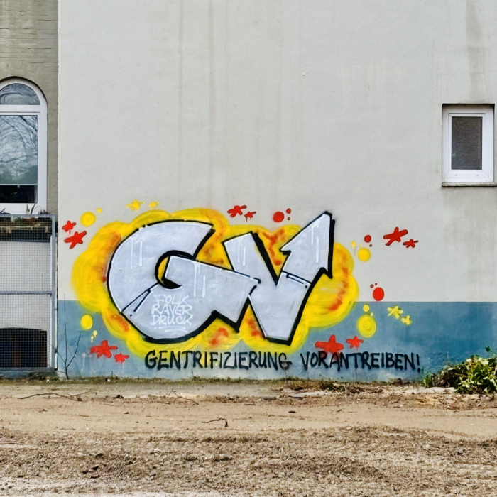 Ein großes Graffiti an einer Mauer hinter einer Brachfläche: "Gentrifizierung vorantreiben!"