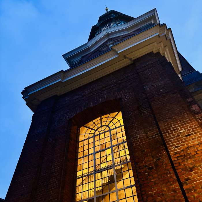 Ein Kirchturm am frühen Abend mit beleuchtetem Fenster, hinter dem man eine Treppe erkennt